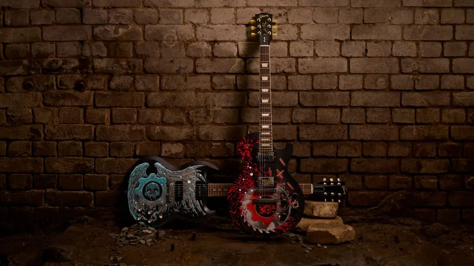 The two Gears of War guitars stood against a brick wall: a black Les Paul painted with a red gear, and a black SG painted with a teal winged skull.